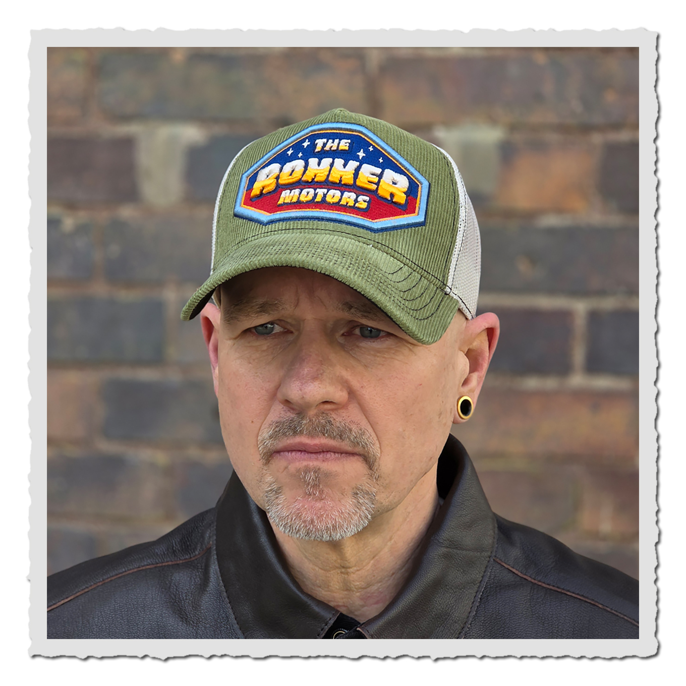 Nahaufnahme einer Person mit moosgrüner und hellgrauer Cap vor Backsteinwand, Gesicht und Jackenkragen sichtbar.